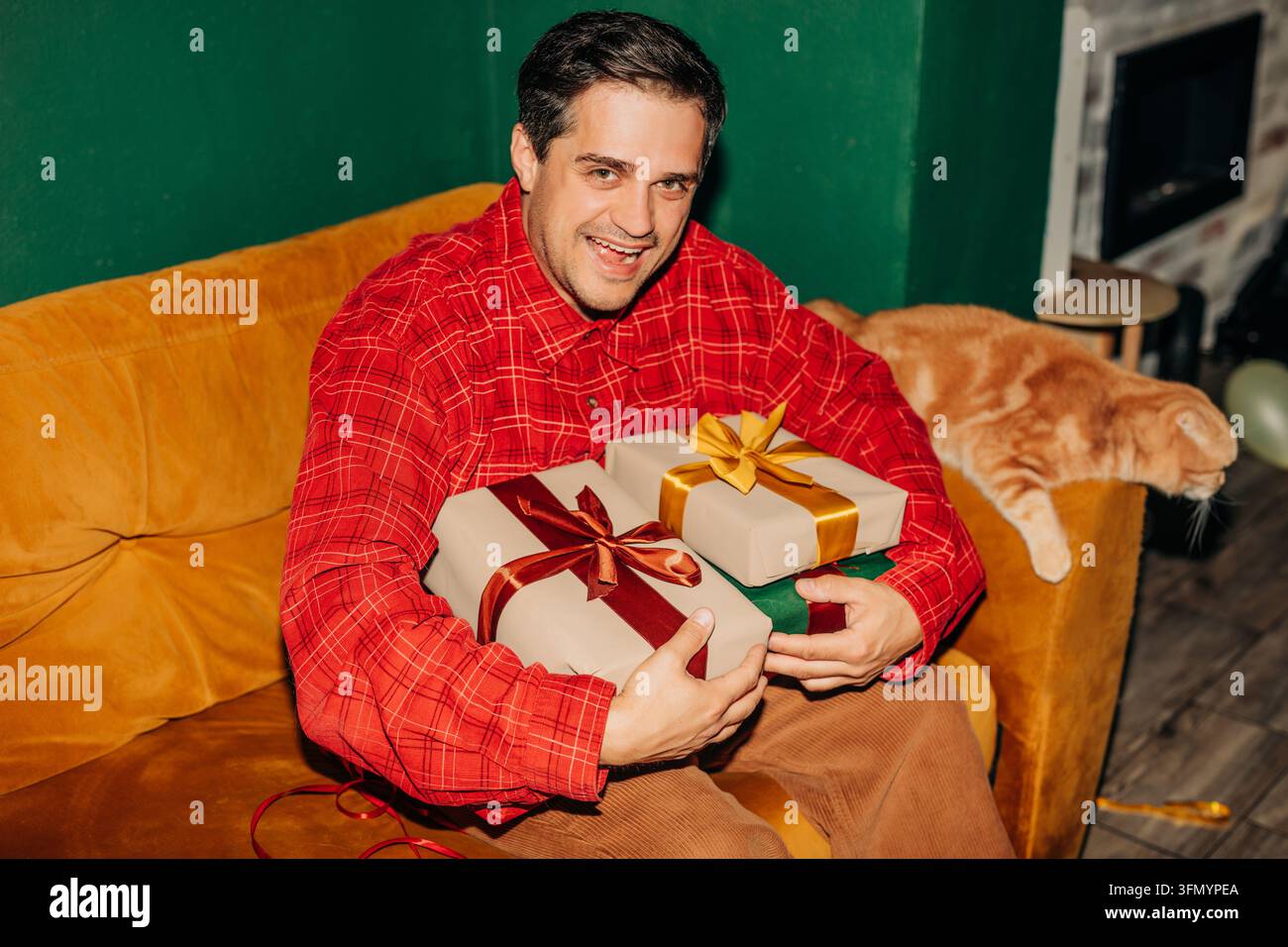 Homme souriant en chemise rouge assis sur un canapé moutarde tenant des cadeaux de Noël emballés avec des rubans tandis qu'un chat orange repose à ses côtés dans une maison confortable Banque D'Images