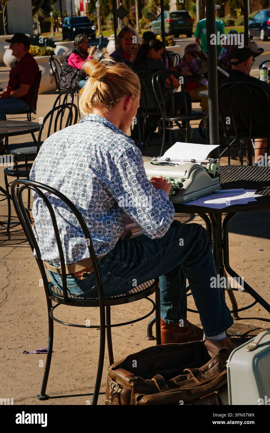 Écrivain tapant sur une machine à écrire manuelle dans un café en terrasse, chemise décontractée et jeans, sac à bandoulière au sol, clients sous les auvents maintenant Banque D'Images