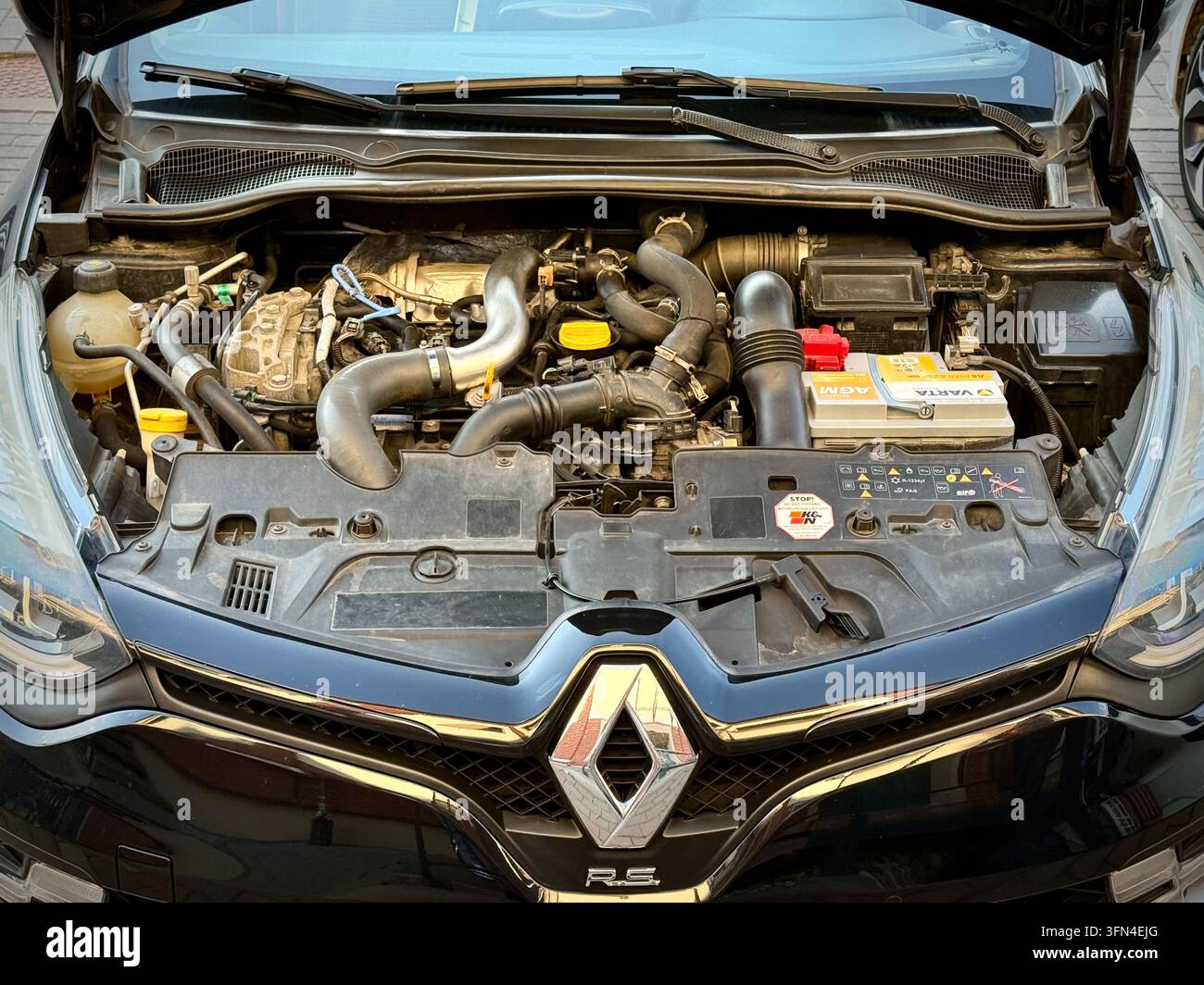 Vue avant du compartiment moteur ouvert d'une Renault Clio RS noire montrant le moteur turbocompressé, la batterie Varta AGM et l'emblème Renault Sport sur la calandre - Image de stock capturée avec un smartphone