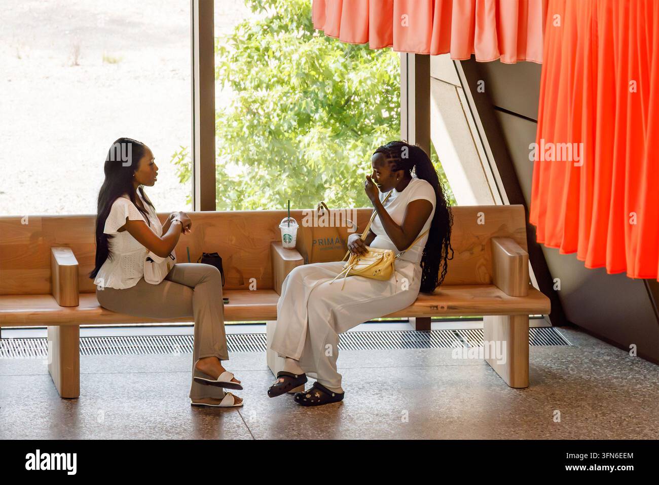 Deux amies noires en blanc assises sur un banc et discutant avec une boisson Starbucks et un sac Primark à l'intérieur près d'une fenêtre du V&A East à Londres Banque D'Images
