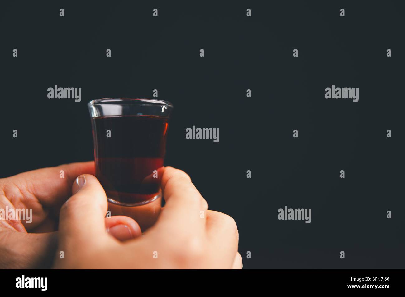 Deux mains tenant un petit verre de boisson de communion rouge foncé contre un fond noir, illustrant un culte chrétien partagé, un souvenir sacré, Banque D'Images