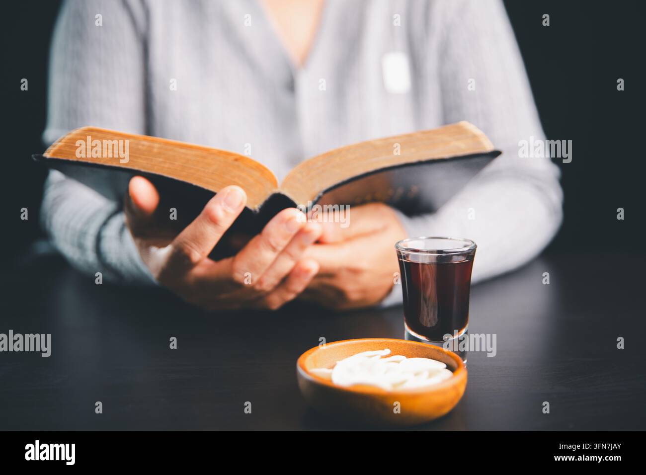 Person lisant un livre ouvert à côté d'hosties et d'une boisson rouge foncé sur une table noire, illustrant le culte chrétien, l'étude des Écritures, la prière Banque D'Images