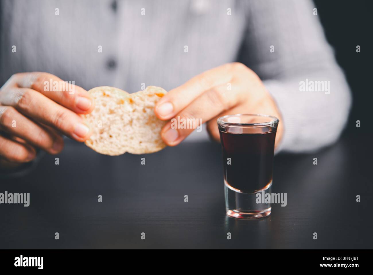 Person brisant du pain à côté d'un petit verre de boisson de communion rouge foncé sur une table noire, représentant le culte chrétien, le souvenir sacré, la foi Banque D'Images