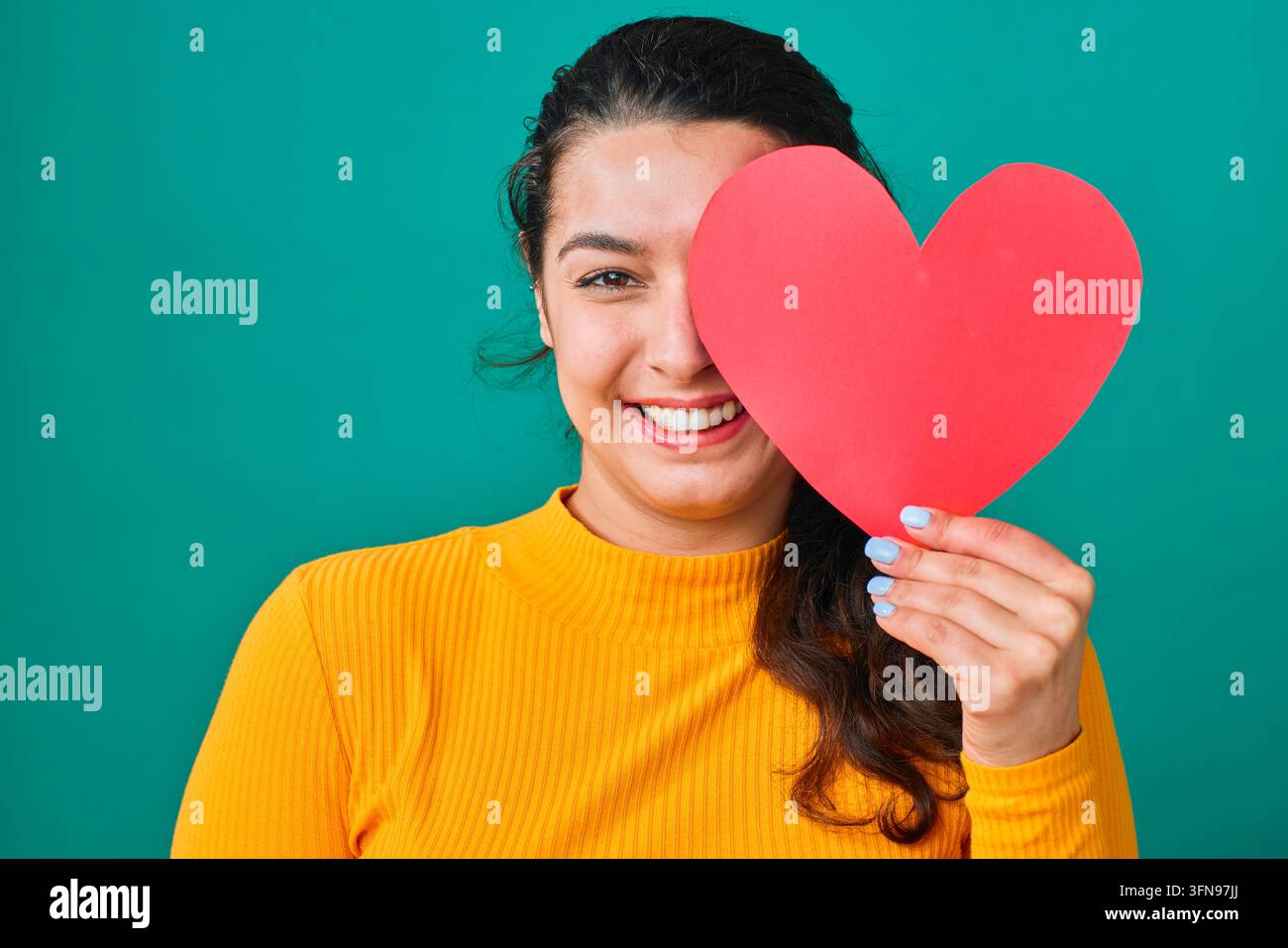 Jeune femme souriante tenant un cœur en papier rouge sur un œil, portrait pour concept d'amour de la Saint-Valentin Banque D'Images