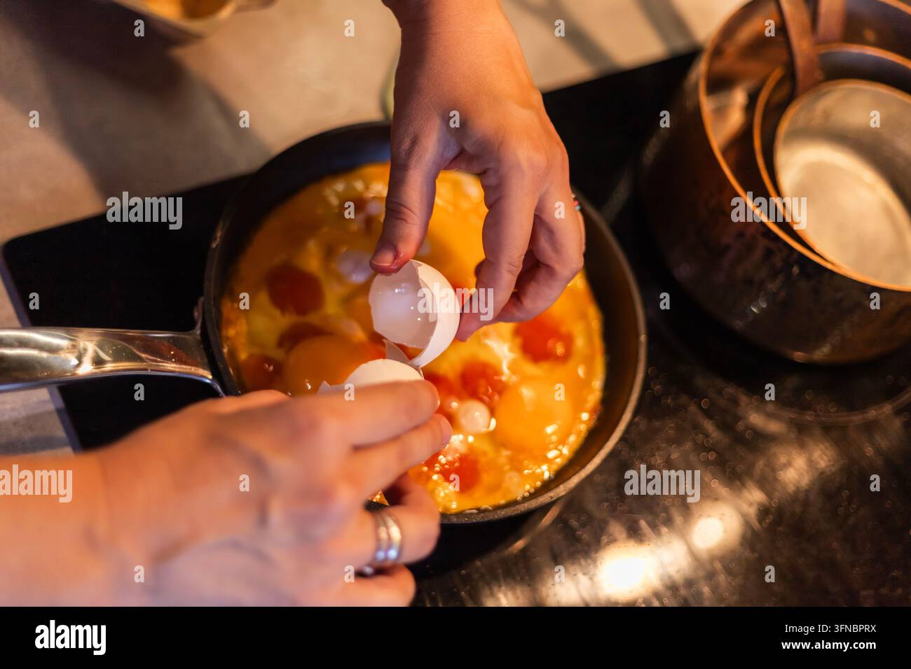 Une paire de mains casse un œuf au-dessus d'une poêle contenant un mélange d'œufs crus. Le gros plan montre les demi-coquilles tenues au-dessus de la poêle sur une cuisinière sombre Banque D'Images