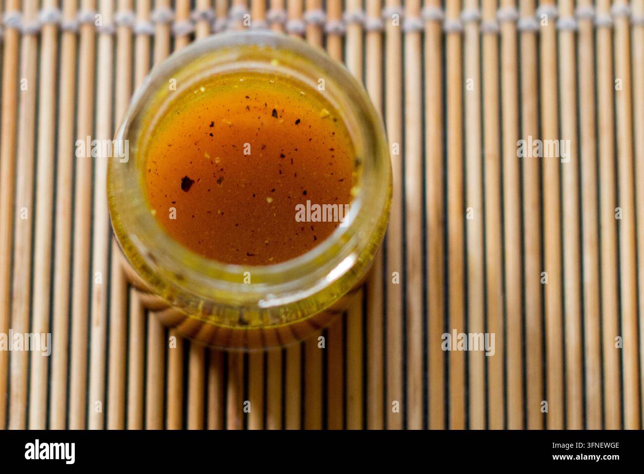 Vue par-dessus du miel doré avec des taches d’épices sombres dans un pot en verre ouvert sur un tapis en bambou, faible profondeur de champ, photographie culinaire en gros plan Banque D'Images