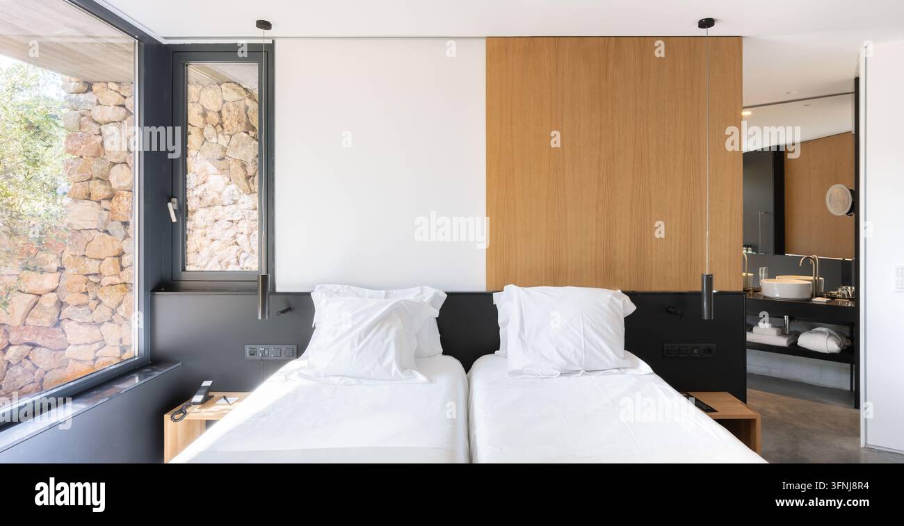 Chambre contemporaine lumineuse dans une villa de luxe en Catalogne, Espagne, avec deux lits jumeaux, des lambris en bois, un mur en pierre, de grandes fenêtres et une salle de bain ouverte Banque D'Images