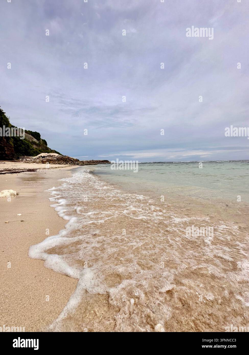 Une douce vague turquoise s'étend sur le sable pâle sous les falaises côtières et les ciels nuageux. - Image de stock capturée avec un smartphone
