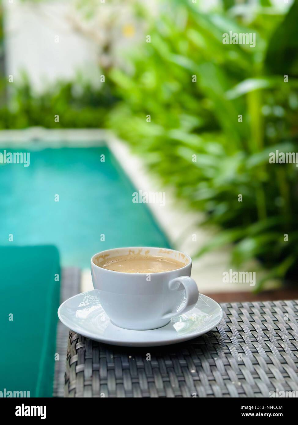 Une tasse de café blanche repose sur des meubles en osier à côté d'une piscine tropicale dans une lumière douce. - Image de stock capturée avec un smartphone