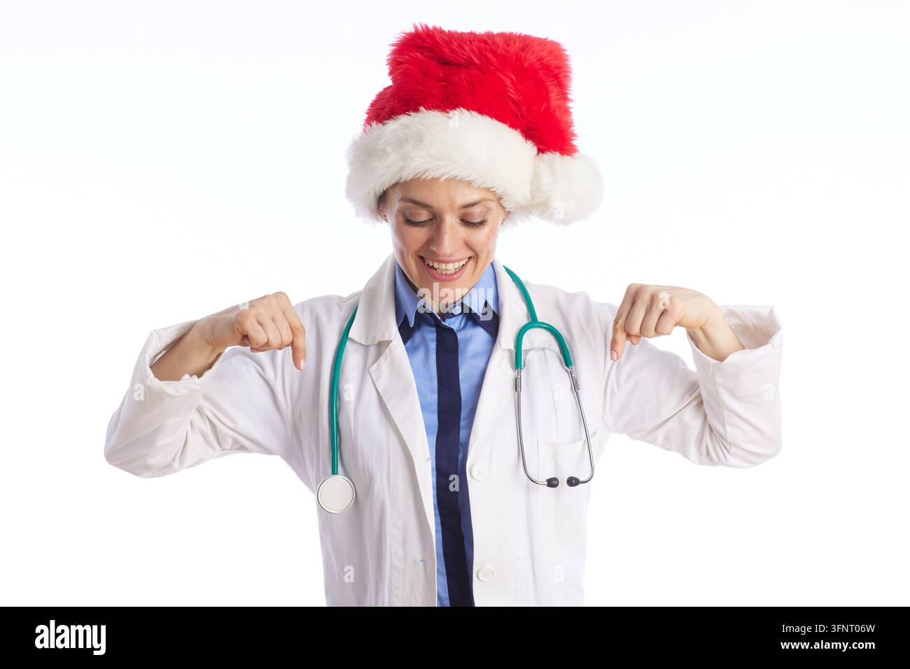 Une jeune médecin avec les yeux fermés et un sourire joyeux porte un chapeau du Père Noël et une blouse de laboratoire, pointant vers ses épaules dans un geste festif des fêtes Banque D'Images