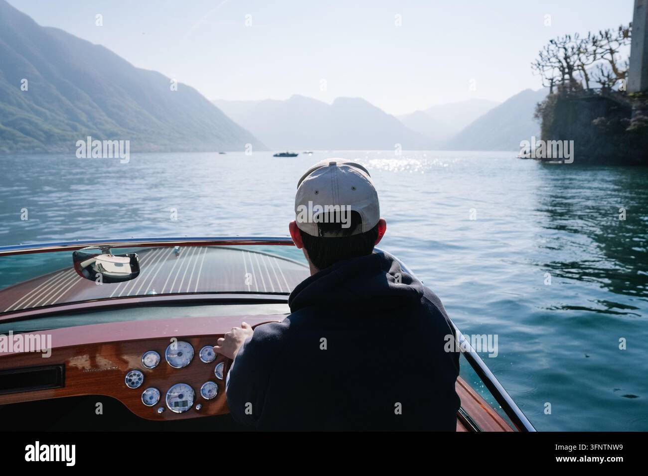 Varenna, Italie - 5 avril 2026 : Homme coiffé d'une casquette blanche dirigeant un bateau à moteur en bois sur le lac de Côme pittoresque Banque D'Images
