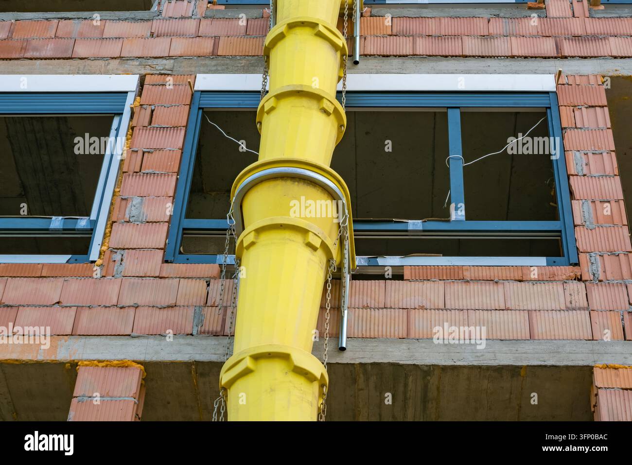 Chute de débris de construction jaune vif sécurisée avec des chaînes contre une façade de briques inachevée avec des sols en béton et un cadre de fenêtre ouvert Banque D'Images