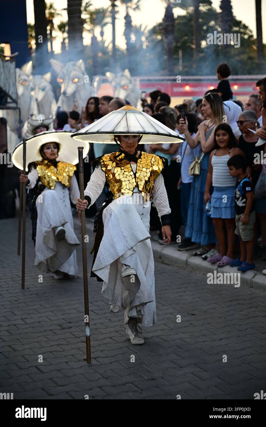 Ville, Pays – 5 octobre 2026 : Des artistes de rue en costumes dorés à miroirs et chapeaux illuminés défilent devant des spectateurs lors d’un festival au crépuscule Banque D'Images