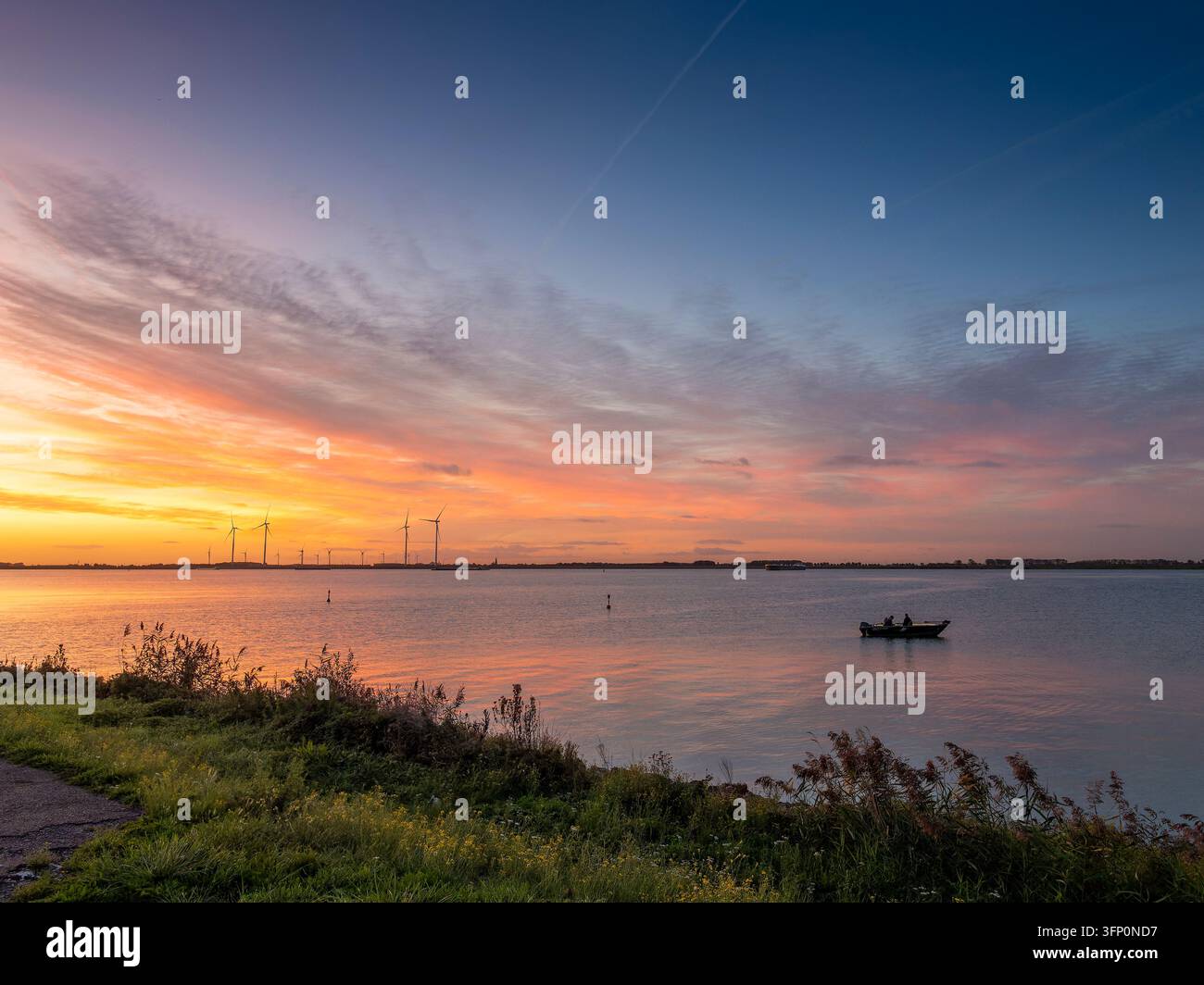 Les couleurs vibrantes du coucher de soleil peignent le ciel au-dessus d'un lac tranquille, reflétant les teintes chaudes tandis qu'un petit bateau avec deux personnes navigue sur les eaux calmes près d'une ferme éolienne à l'horizon Banque D'Images