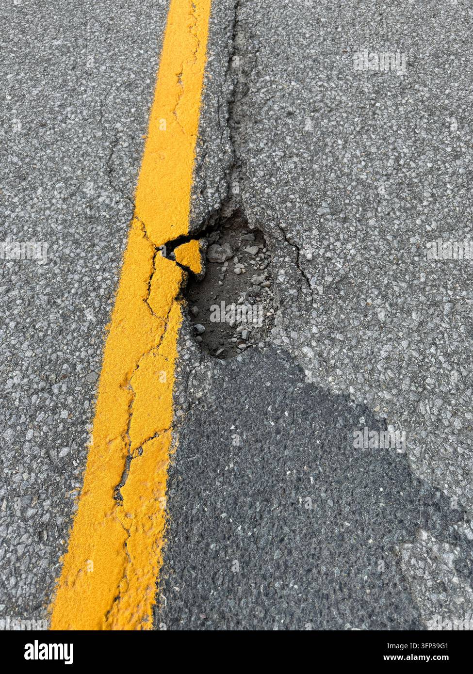 Nid de poule et fissure dans la surface en asphalte adjacente à un séparateur de voie peint en jaune sur une route pavée. Banque D'Images