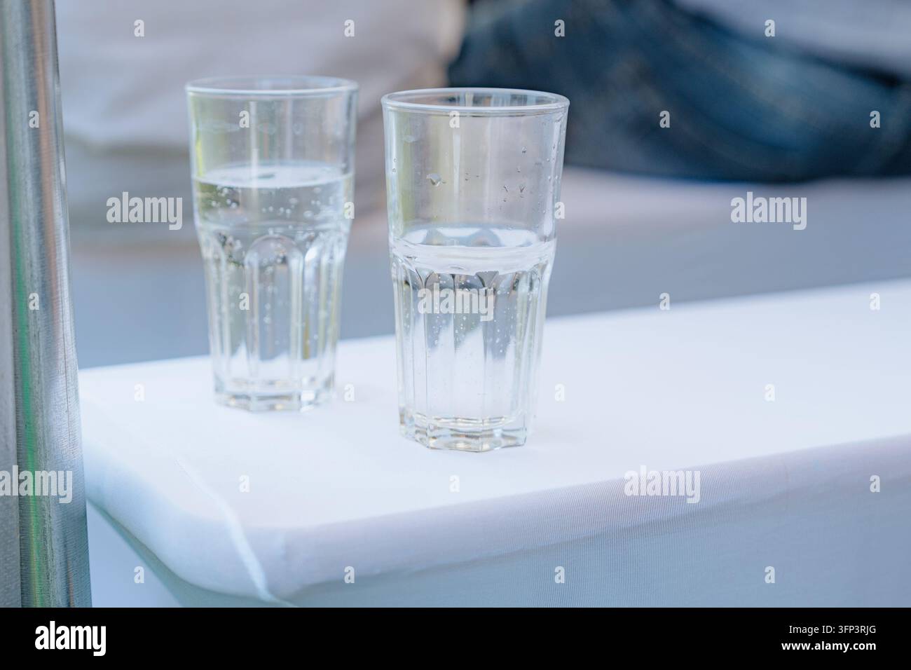 Deux verres d'eau sont posés sur une surface recouverte d'un tissu blanc dans un cadre extérieur avec un poteau métallique, capturés de près, légèrement surélevés Banque D'Images