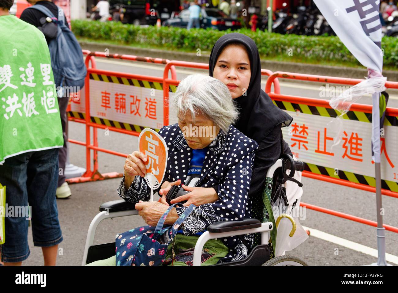 Taipei, Taïwan - 03 oct. 2026 : Une aide-soignante migrante portant un hijab s'occupe d'une femme âgée en fauteuil roulant tenant un éventail à côté des barrières "No Entry". Banque D'Images