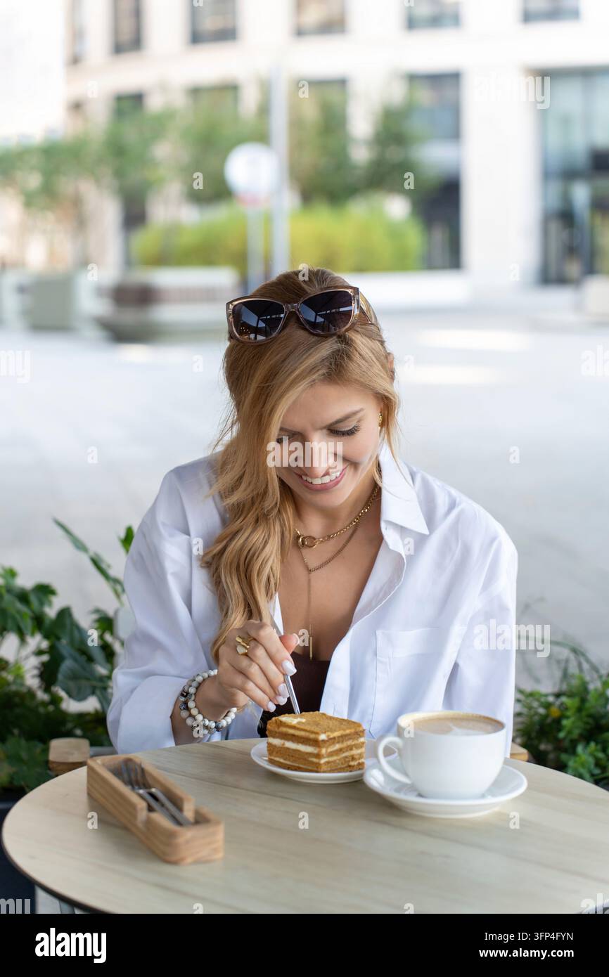 Une scène calme de café en plein air avec une femme portant des bijoux et des lunettes de soleil, savourant un gâteau à étages. Élégance féminine et charme urbain Banque D'Images