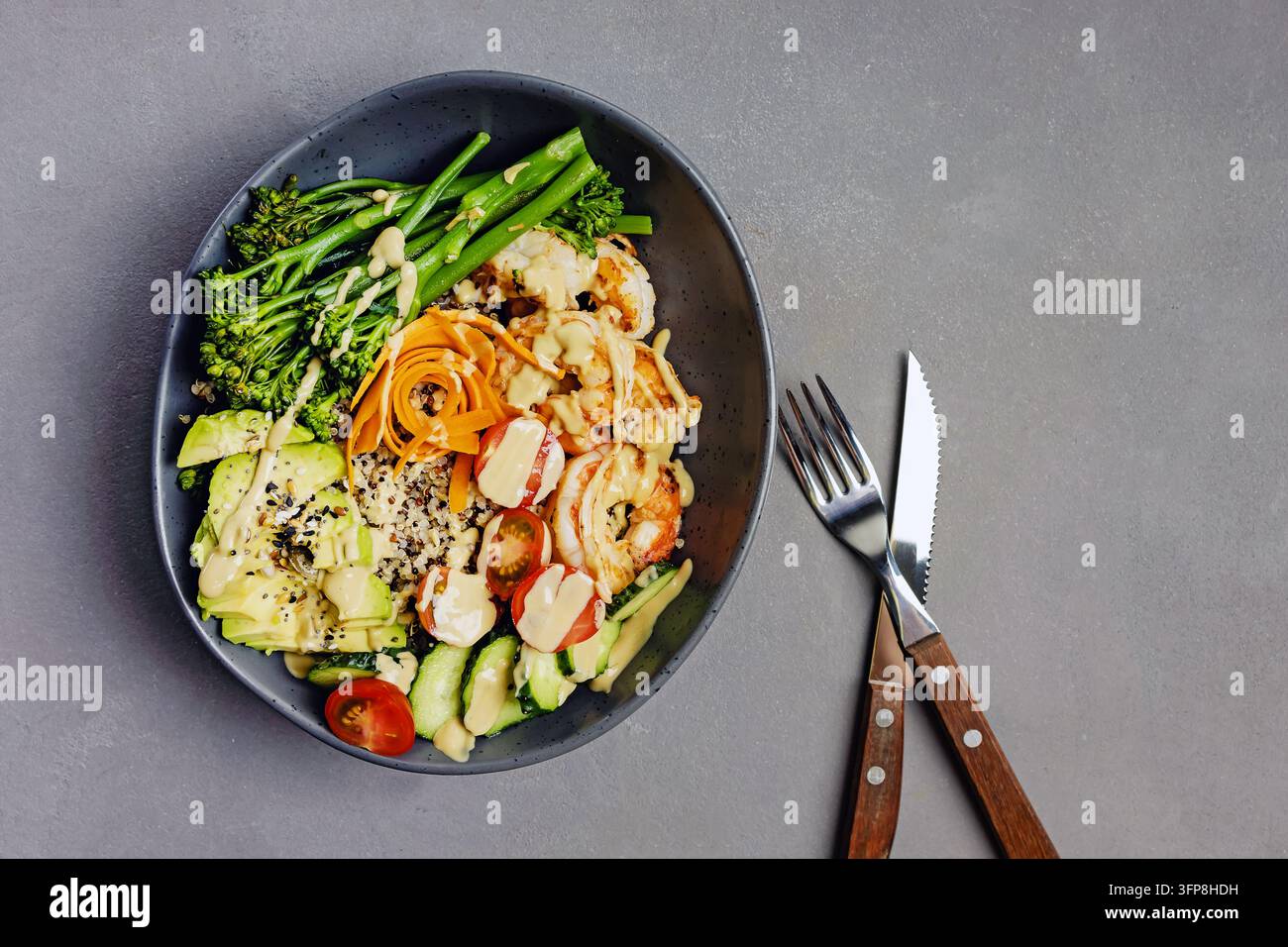 Bol sain de crevettes grillées, quinoa, avocat, broccolini, concombre, tomates et rubans de carotte avec une sauce crémeuse, fourchette et couteau sur gris. Banque D'Images