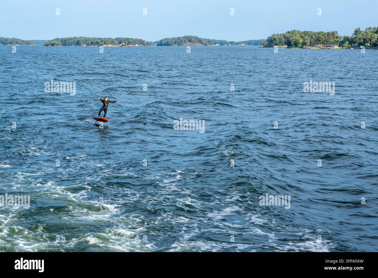 Archipel de Stockholm, Suède ; 13 août 2026 : Surfeur en combinaison de plongée chevauchant une planche à voile à hydrojet propulsée par les vagues de sillage créées par un bateau qui passe Banque D'Images