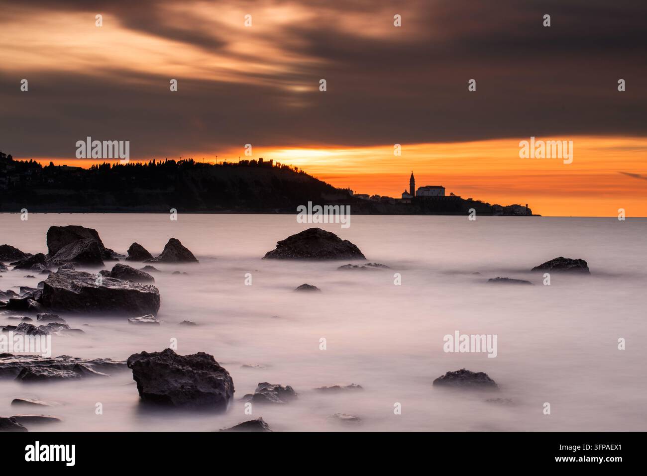 Vue de la silhouette de l'église Saint-Georges à Piran au coucher du soleil, avec une côte rocheuse au premier plan et une mer calme sous un ciel orange dramatique Banque D'Images