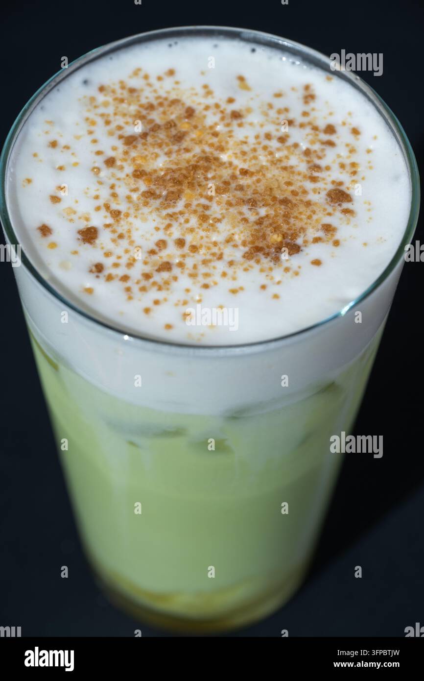 Un grand verre de latte glacé au thé vert matcha rafraîchissant, surmonté d'une épaisse mousse de lait blanche et saupoudré de poudre d'épices aromatiques sur fond noir Banque D'Images