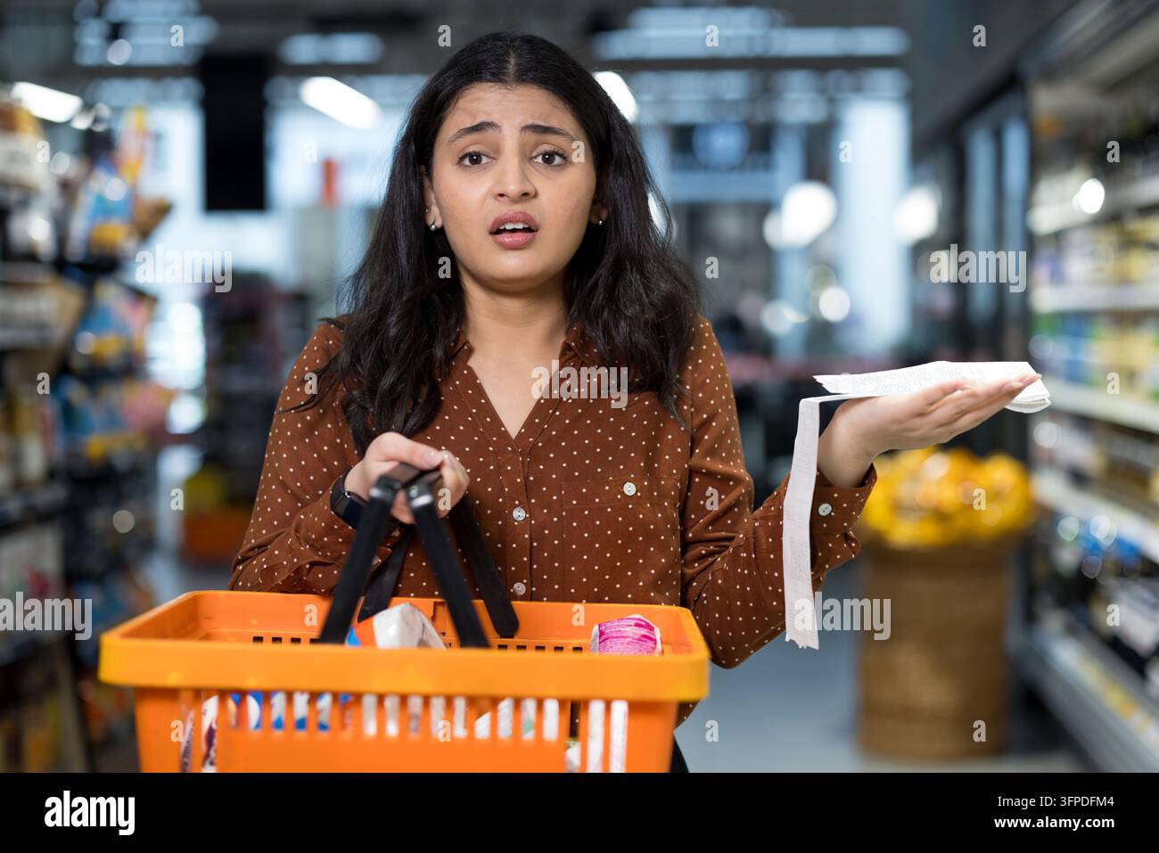 Femme debout dans un rayon de supermarché tenant un panier et un long ticket de caisse, l’air choquée et inquiète face à la hausse du coût des produits d’épicerie et à l’inflation Banque D'Images