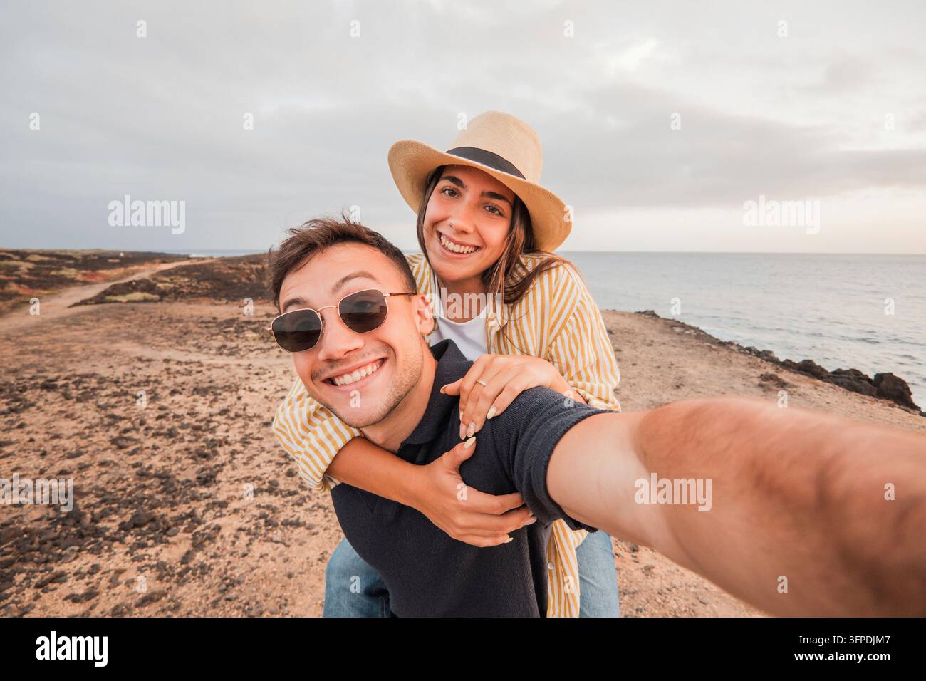 Jeune homme donnant un tour sur ses épaules à sa petite amie tout en souriant à l'appareil, capturant un selfie joyeux ensemble pendant leurs vacances au bord de l'océan au coucher du soleil Banque D'Images