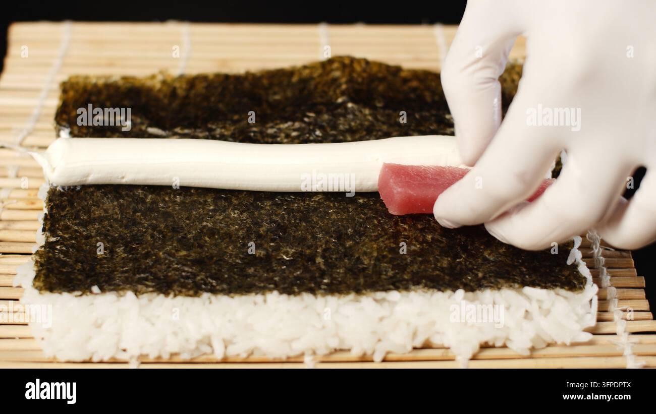 Main gantée d'un chef posant du thon frais sur des algues nori avec du fromage frais et du riz à sushi sur un tapis en bambou Banque D'Images