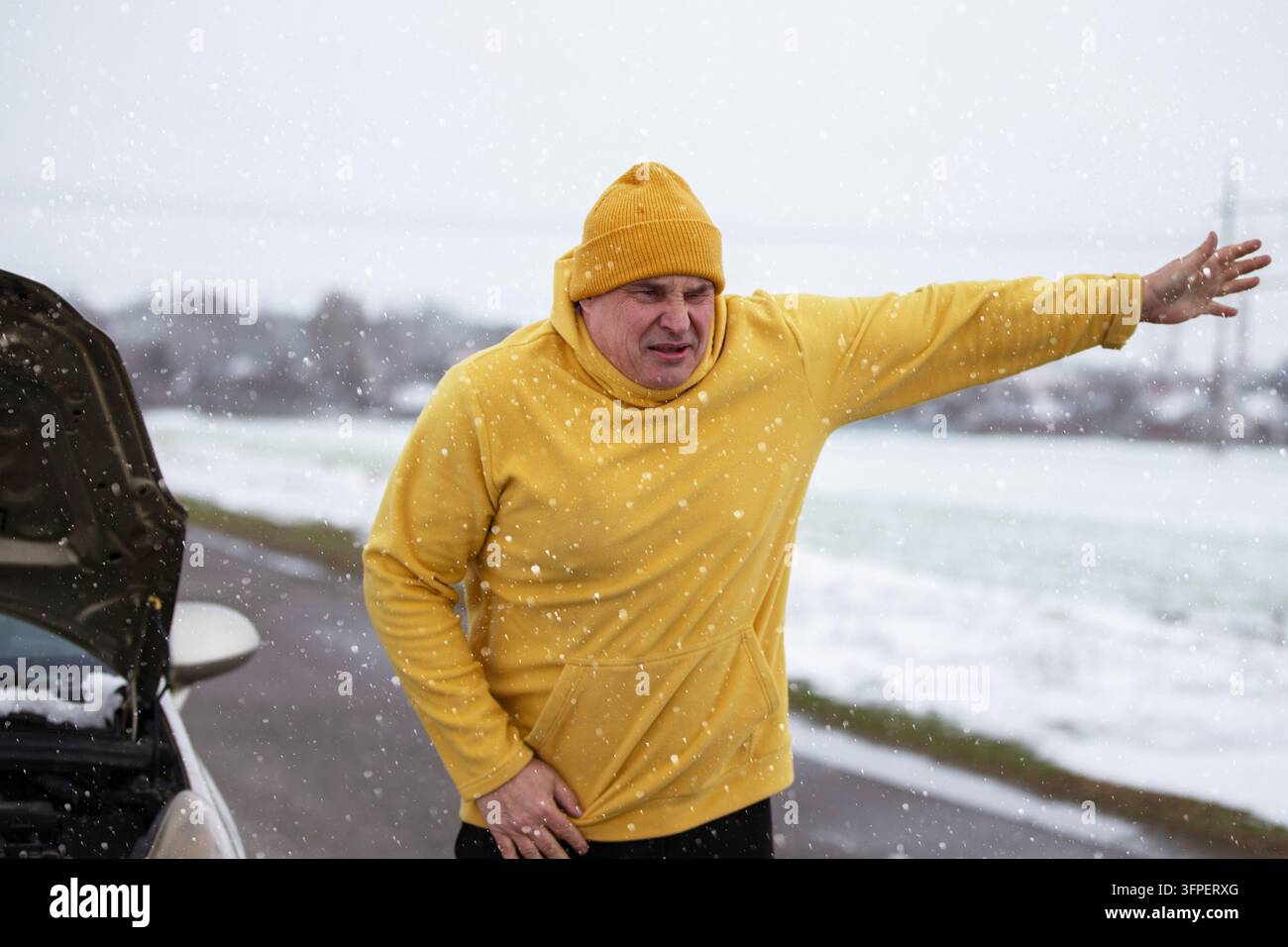 Un homme dans un sweat-shirt jaune vote sur la route à côté d'une voiture en panne. Il a l'air contrarié, peut-être à cause de problèmes avec sa voiture par temps hivernal. Banque D'Images
