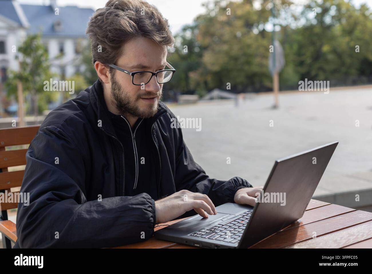 Un homme barbu portant des lunettes et une veste noire tape sur un ordinateur portable alors qu'il est assis à une table en bois dans un espace public extérieur. Arbres, piétons, pavé Banque D'Images