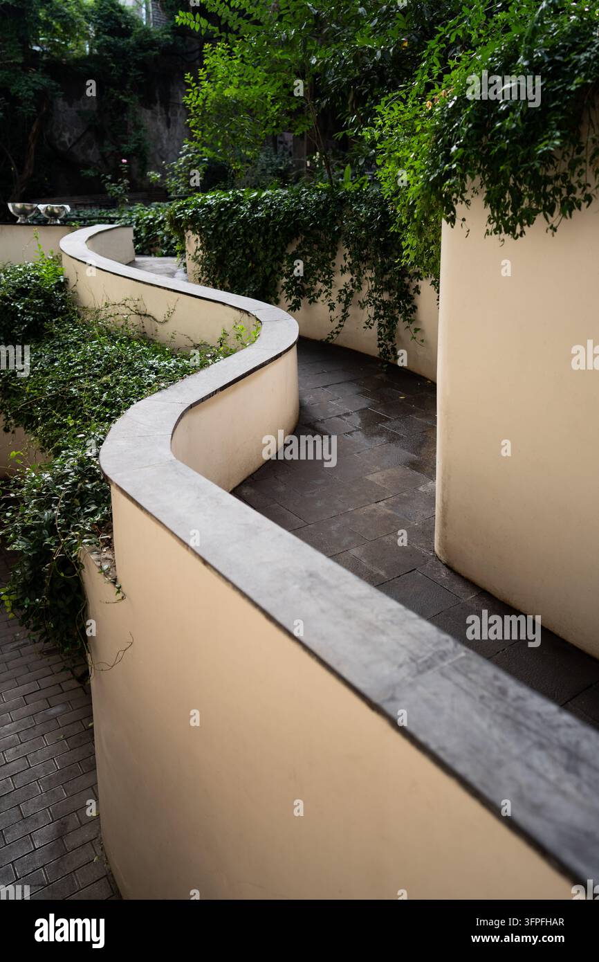 Allée piétonne incurvée bordée de murs crème et d'une végétation dense dans un jardin urbain paysagé Banque D'Images