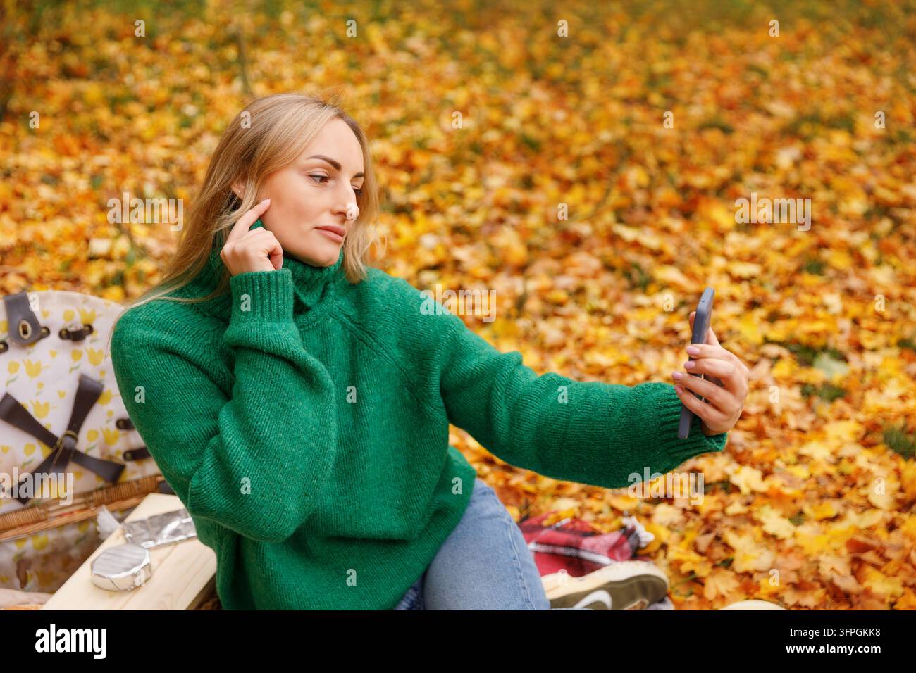 Session photo automnale moderne dans le parc : jeune femme en pull vert ajustant un écouteur sans fil et prenant un selfie sur smartphone Banque D'Images