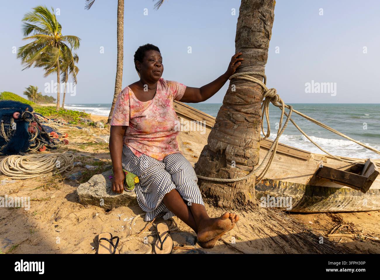 Une femme est assise à côté d'un bateau de pêche en bois sous un palmier sur la plage, région centrale, Elmina, Ghana Banque D'Images