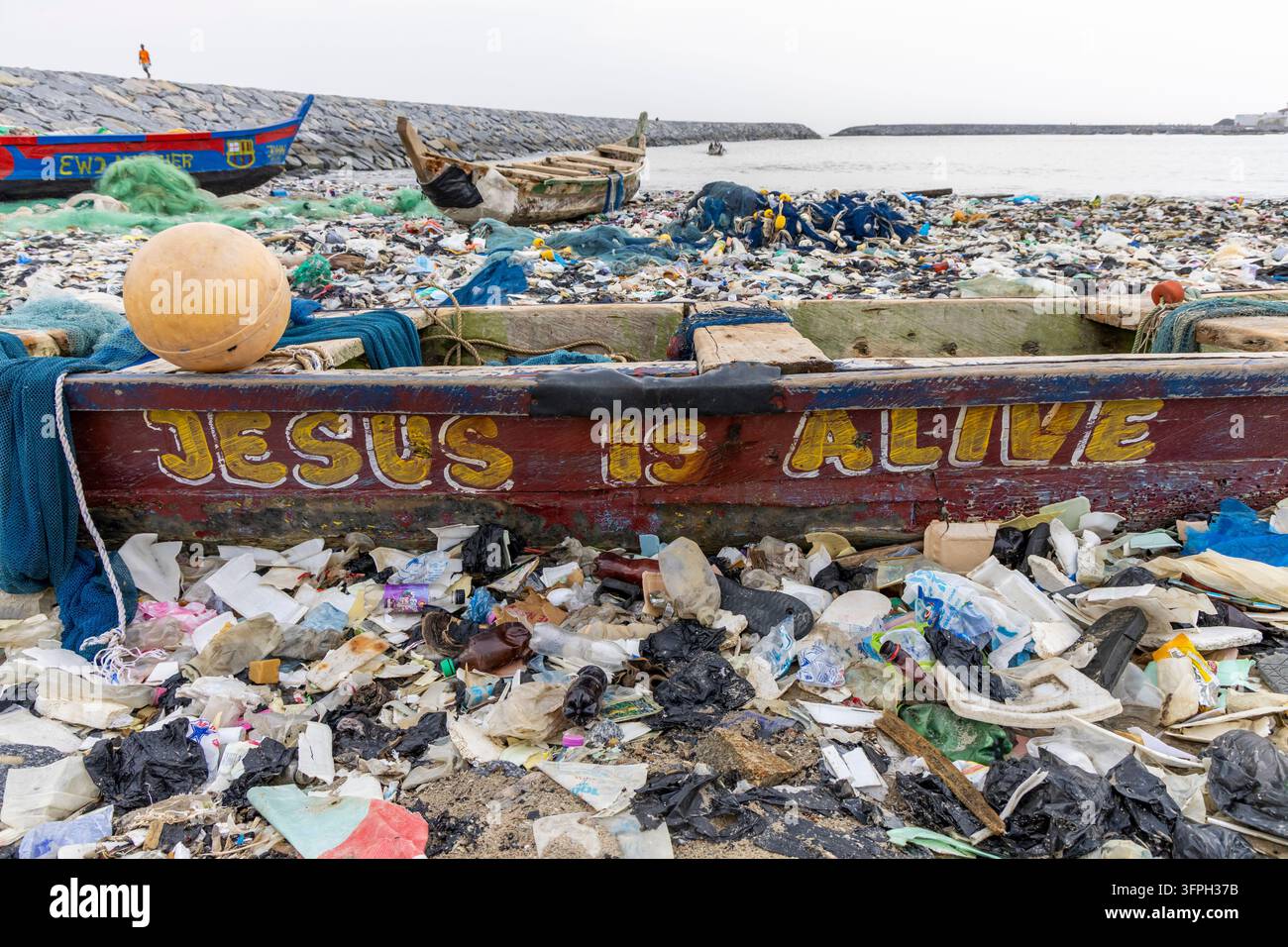 Vue rapprochée d'un bateau de pêche en bois peint avec les mots Jésus est vivant avec un bateau sur une plage couverte de déchets plastiques, Cape Coast, Ghana Banque D'Images