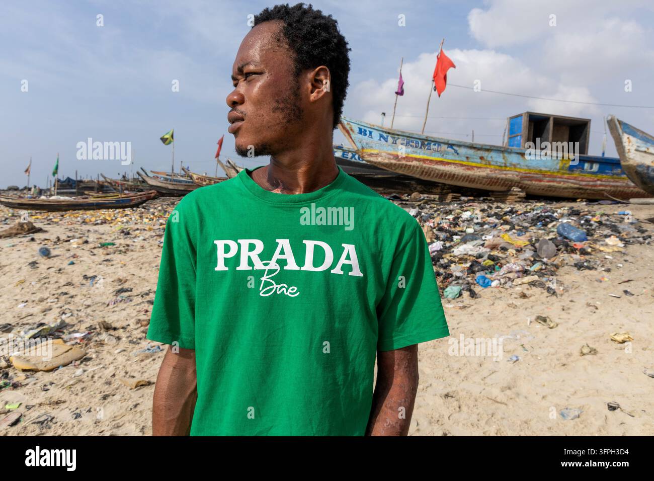 Un homme portant un t-shirt vert se tient sur une plage de sable jonchée de déchets plastiques avec des bateaux de pêche traditionnels amarrés derrière lui, Région de Greater Accra, Acc Banque D'Images