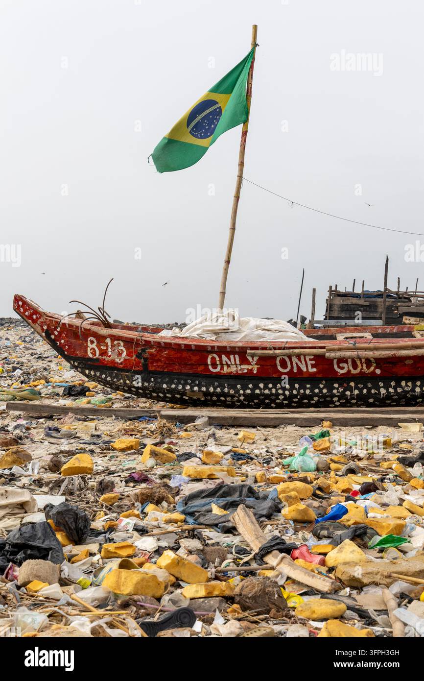 Un bateau de pêche en bois peint arborant un drapeau brésilien repose sur une plage couverte de déchets plastiques, région de Greater Accra, Accra, Ghana Banque D'Images