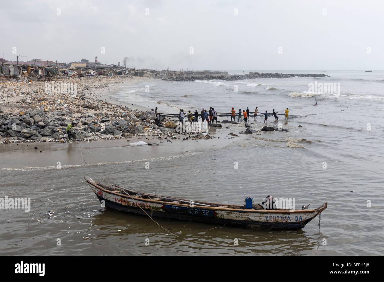 Pêcheurs tirant un filet de pêche sur une plage polluée avec un bateau en bois au premier plan, région de Greater Accra, Accra, Ghana Banque D'Images
