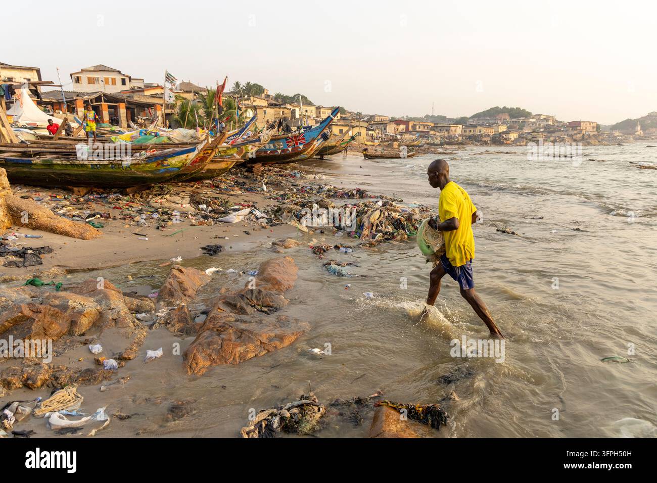 Homme en chemise jaune portant un filet de pêche en marchant dans des eaux peu profondes, Région centrale, Cape Coast, Ghana Banque D'Images