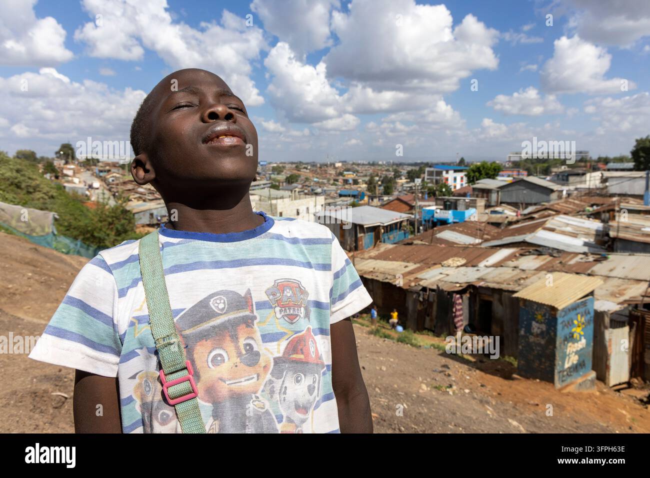 Garçon portant un t-shirt rayé Paw Patrol, la tête penchée en arrière les yeux fermés au-dessus des toits de la zone de Kibera, comté de Nairobi, Nairobi, Kenya Banque D'Images