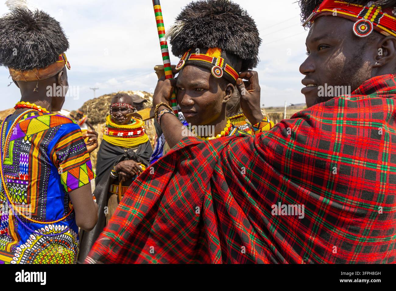 Homme de la tribu Turkana portant un châle à carreaux rouges aidant un jeune homme à ajuster son couvre-chef à plumes noires et son bandeau perlé au Festival culturel du lac Turkana Banque D'Images