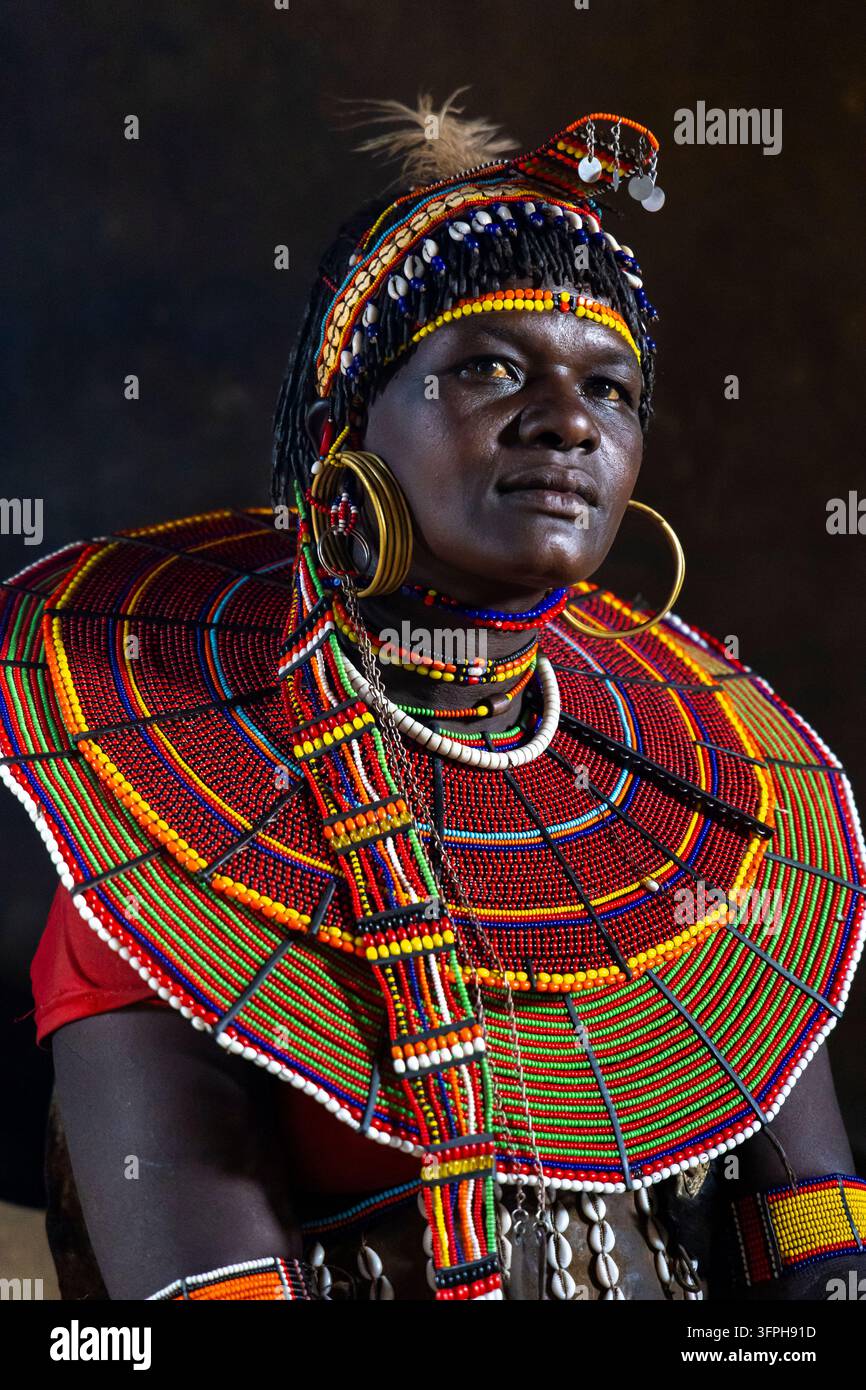 Portrait d'une femme de la tribu Pokot dans une hutte sombre portant un coiffeur traditionnel en plumes, des colliers de coquilles de cauri, des boucles d'oreilles en laiton et un énorme collier de perles, regardant vers le haut Banque D'Images