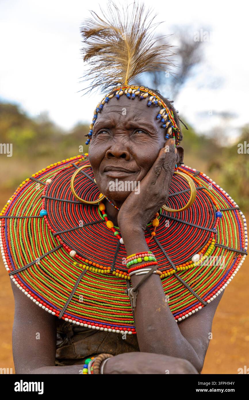 Femme de la tribu Pokot avec un bandeau à plumes et coquilles de cowrie et un large collier de perles, regardant vers le haut avec la main sur la joue, Narok, Chepilat, Kenya Banque D'Images