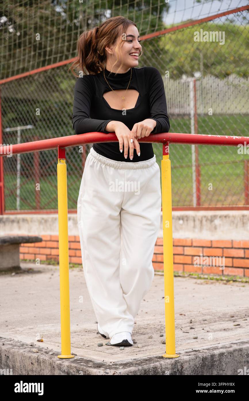 Jeune femme souriant en plein air, vêtue d'un haut noir et d'un pantalon blanc, s'appuyant de manière décontractée sur une rambarde colorée dans un terrain de sport, exprimant le bonheur, y Banque D'Images