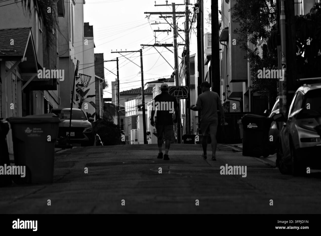 Deux hommes descendent une rue étroite au crépuscule. Les silhouettes montrent une promenade décontractée, une ambiance urbaine paisible. Les bâtiments bordent la route, des voitures garées flanquent le chemin. La scène suggère Banque D'Images