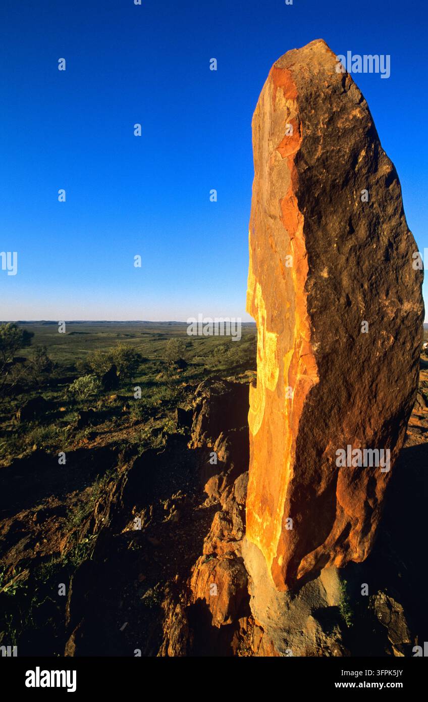 Monolithe de pierre érodé éclairé par le soleil bas sur une crête rocheuse, avec des broussailles s'étendant jusqu'à l'horizon sous un ciel bleu clair dans un pays aride et reculé. Banque D'Images
