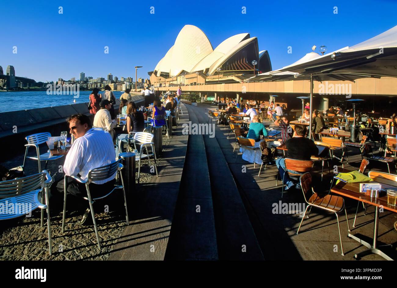 Les clients sont assis à des tables au bord de l'eau sur la promenade de Bennelong Point sous les voiles de l'Opéra, avec une vue sur le port de Sydney et le pilier du pont. Banque D'Images