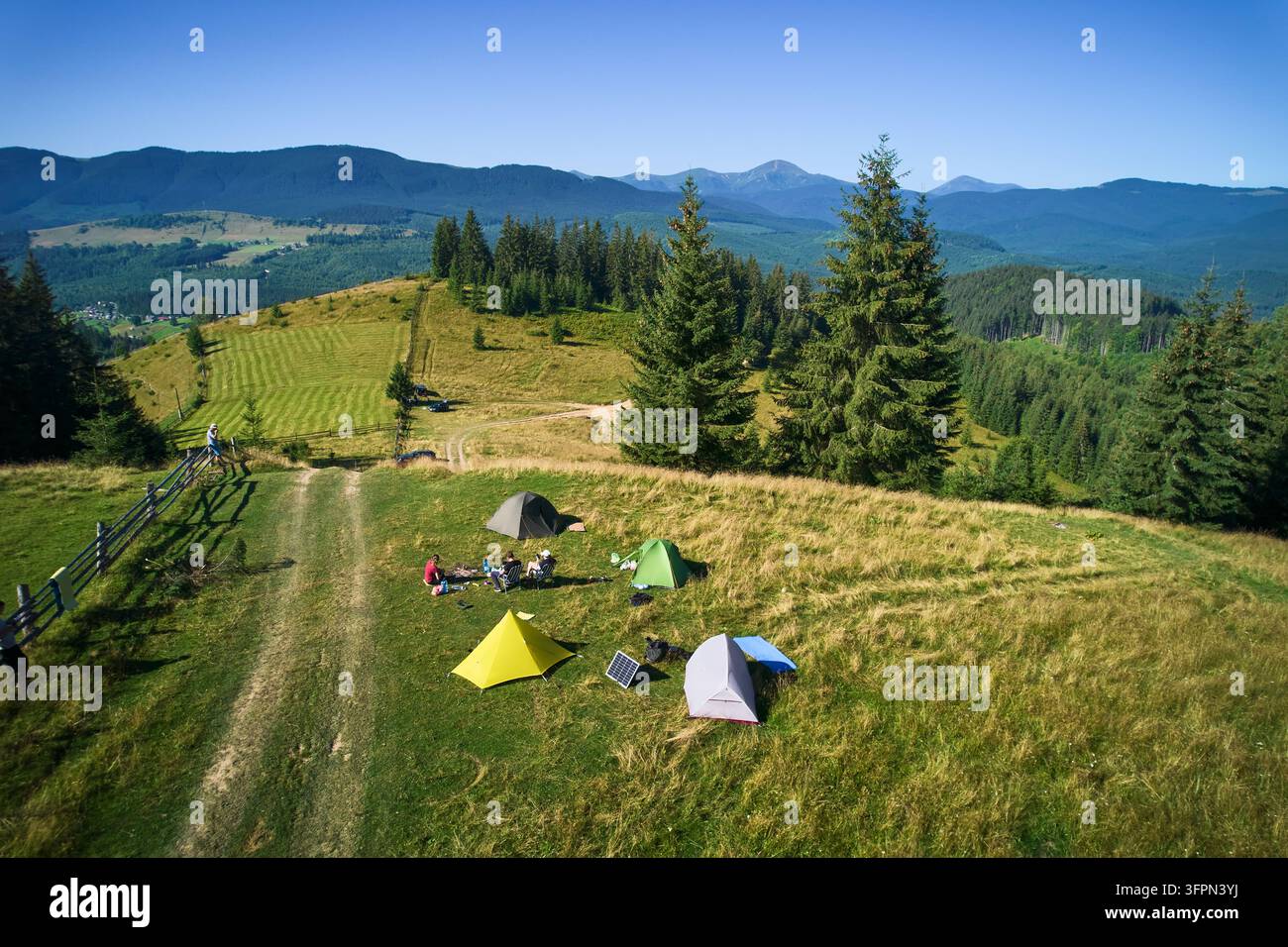 Camping vue aérienne dans les montagnes, avec plusieurs tentes colorées sur clairière herbeuse. Les campeurs se détendent sous un ciel bleu clair, entourés de hauts conifères et de vues panoramiques sur des sommets lointains. Banque D'Images