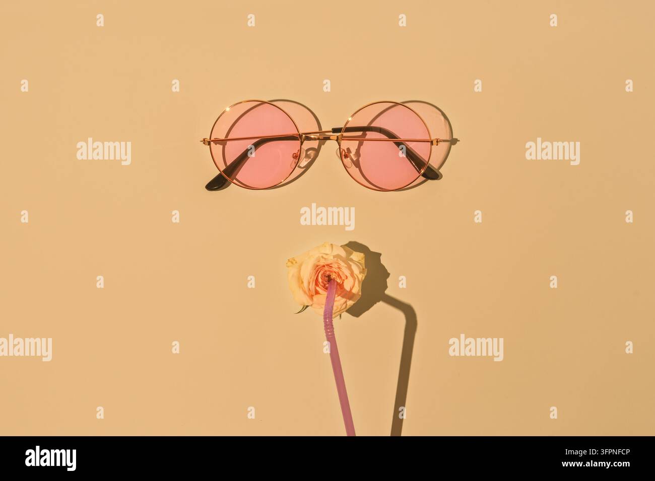 Nature morte minimaliste élégante avec des lunettes de soleil roses, une rose et de la paille projetant des ombres sur un fond beige chaud sous une lumière solaire vive Banque D'Images