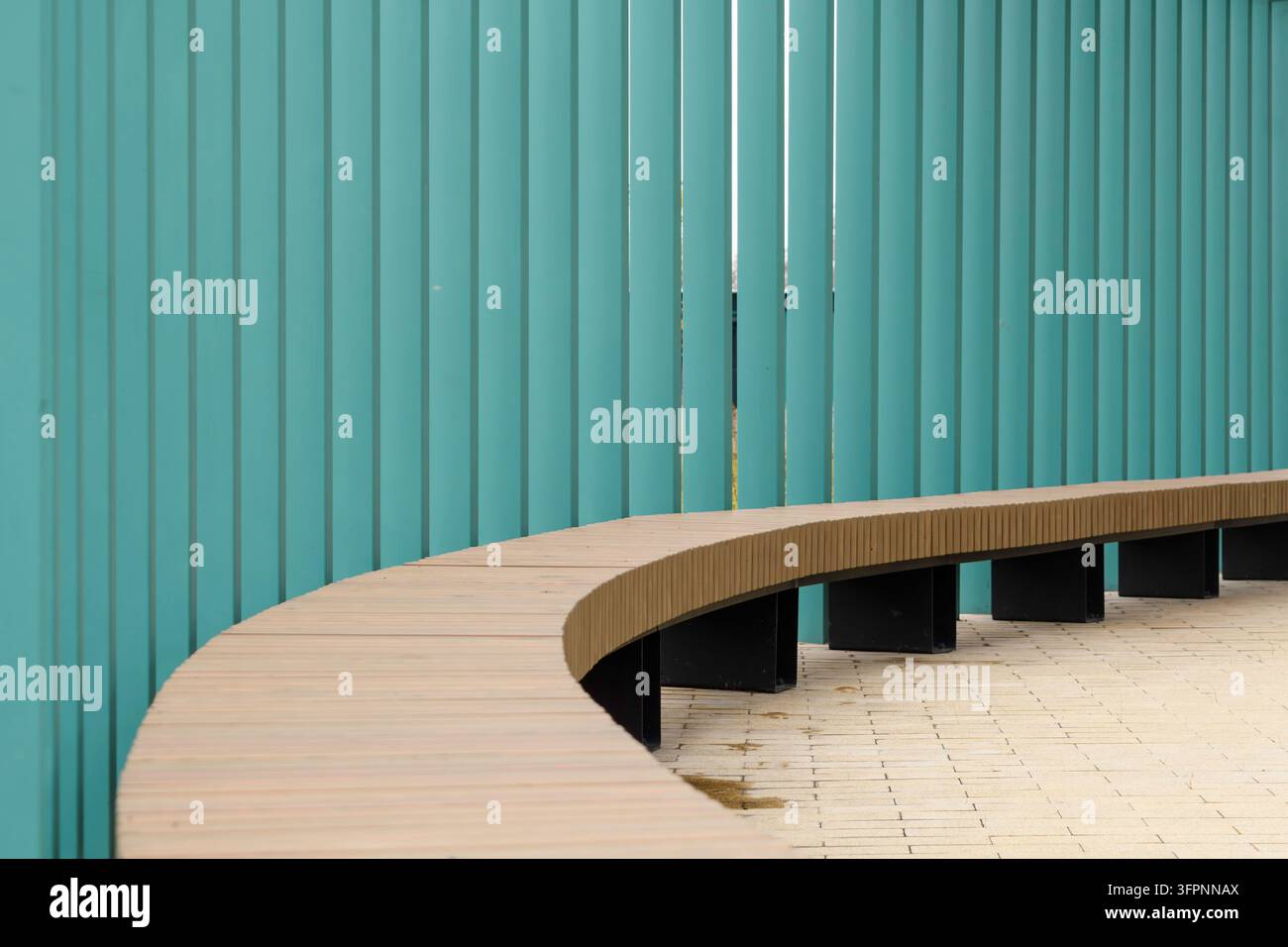 Un banc en bois incurvé contre des lamelles verticales turquoise crée un espace urbain moderne aux lignes épurées, au minimalisme et au rythme géométrique. Banque D'Images